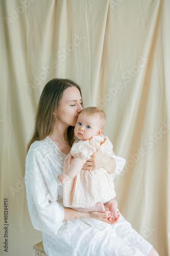 Young beautiful mother in a white dress is holding her little blue-eyed daughter of 5 months in her arms. Motherhood.
