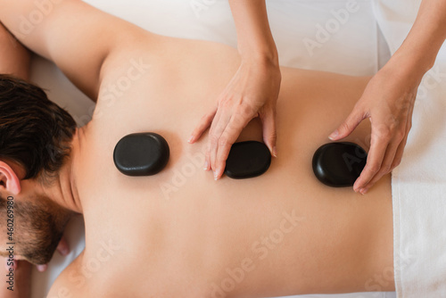 Cropped view of masseur putting hot stones on back of man in spa center