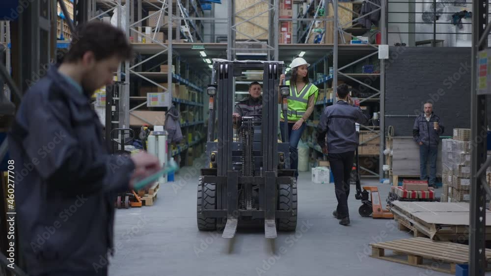 Wide shot forklift driving in warehouse in slow motion with serious concentrated freight agent in hard hat standing on machinery talking with driver pointing around. Professional people working Wide shot forklift driving in warehouse in slow motion with serious concentrated freight agent in hard hat standing on machinery talking with driver pointing around. Professional people working