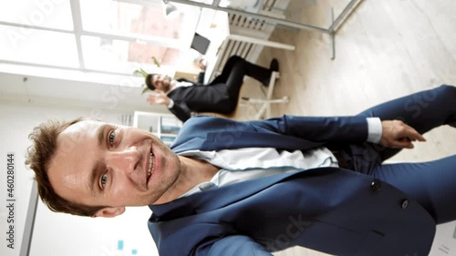 POV vertical of middle-aged Caucasian male office worker filming him and colleague sitting by desk in bright office, talking, smiling and waving on camera for video blog photo