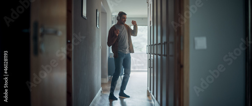 Portrait of a Happy Young Adult Man Listening to Music on Headphones Playing from His Smartphone at Home. Stylish Male in Casual Clothes is Carefree and Dancing in Corridor in His Apartment.