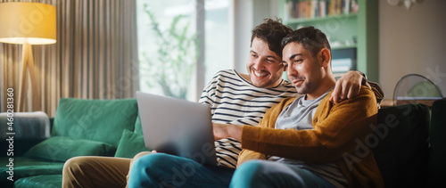 Portrait of Gentle Gay Couple Using Laptop Computer, while Sitting on a Couch in Cozy Stylish Apartment. Adult Boyfriends Online Shopping on Internet, Watching Funny Videos on Streaming Service. photo