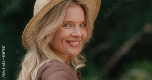 Happy Beautiful Middle-Aged Woman Walking at Park while turning to the camera. Amazing Caucasian Lady with Blond Hair and Expressive Blue Eyes wears Straw Hat. Natural Appearance. Adult Female.