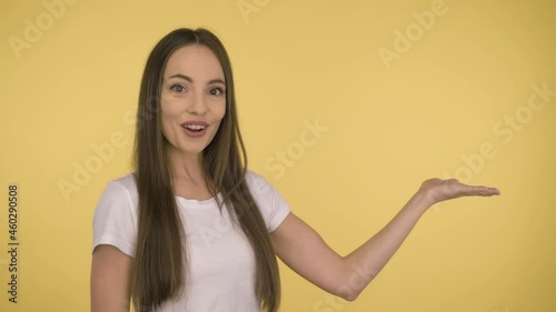 Attractive smiling woman 35 years old in a white basic t-shirt on a yellow background. Shows the offer on the palm.
