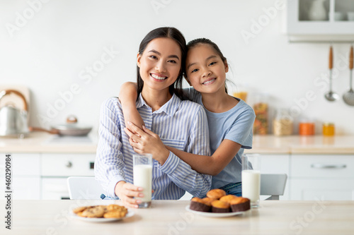 Loving korean mother and her daughter enjoying fresh homemade cookies, drinking milk, sitting in kitchen interior