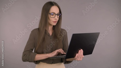 Attractive smiling business woman for 30 years in a dark green office shirt and in glasses. Woman working on a laptop while standing on a gray background.