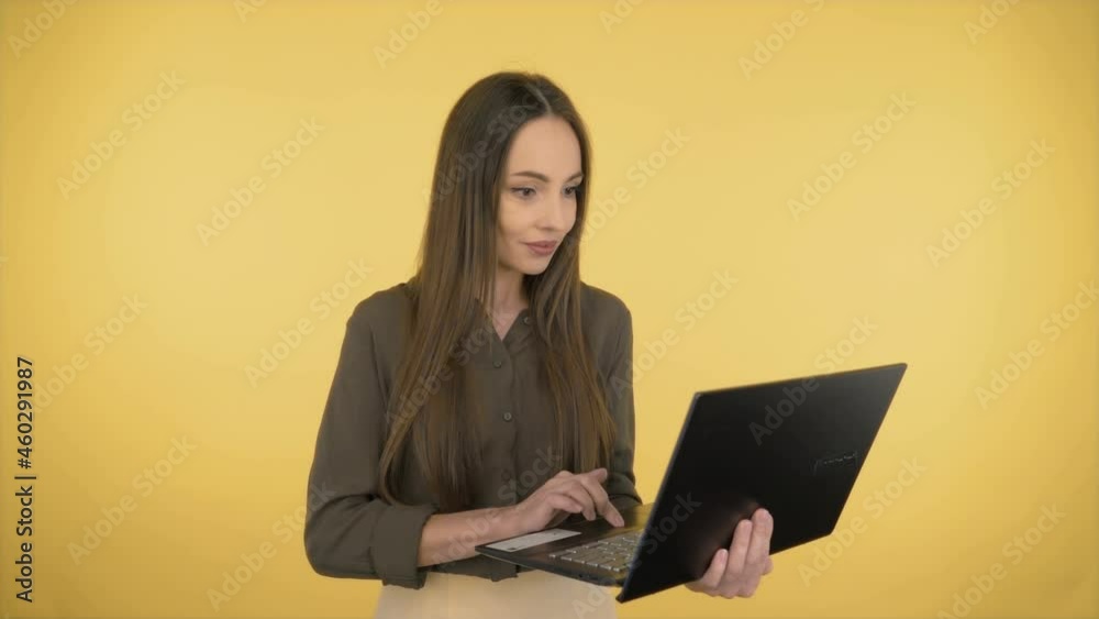 Attractive smiling business woman in a dark green office shirt. Woman working on a laptop while standing on yellow background.