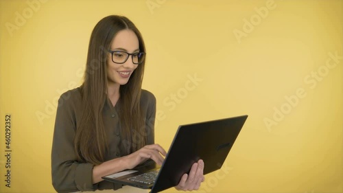 Attractive smiling business woman in a dark green office shirt and black glasses. Woman working on a laptop while standing on yellow background.