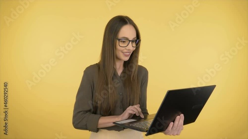 Attractive smiling business woman in a dark green office shirt and black glasses. Woman working on a laptop while standing on yellow background.