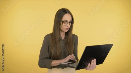 Attractive business woman in a dark green office shirt and black glasses. Woman working on a laptop while standing on yellow background.