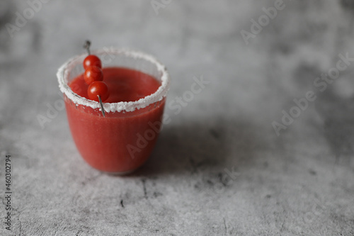 Glass of fresh red tomato juice with salt and metal stick with candy tomatoes on grey background.