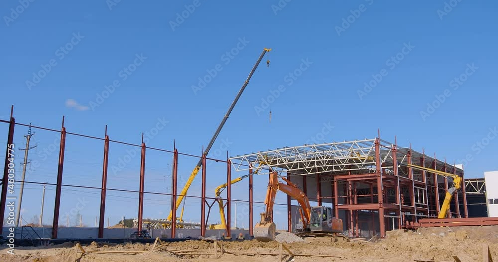 At construction site, construction machinery works next to a factory workshop under construction -the excavator digs the ground, the car lift lifted the builders to the top of the structure At construction site, construction machinery works next to a factory workshop under construction -the excavator digs the ground, the car lift lifted the builders to the top of the structure
