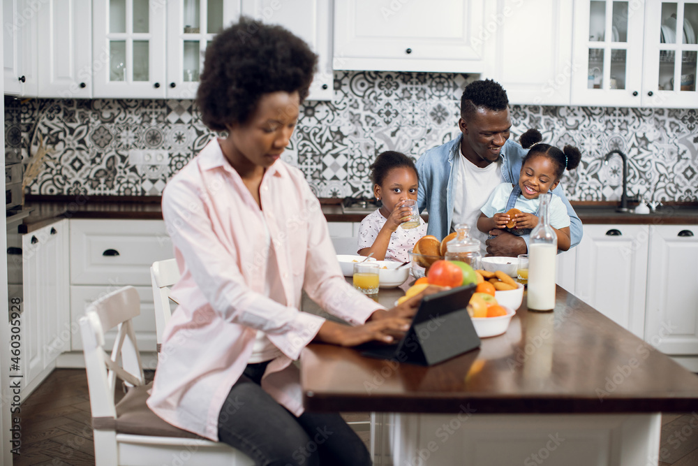 Two pretty girls playing with father during breakfast on kitchen while their african mother working on digital tablet. Concept of family, technology and freelance. Two pretty girls playing with father during breakfast on kitchen while their african mother working on digital tablet. Concept of family, technology and freelance.