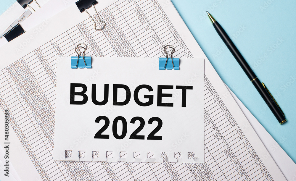 On a light blue background, documents, a pen and a sheet of paper on blue paper clips with the text BUDGET 2022. Business concept. On a light blue background, documents, a pen and a sheet of paper on blue paper clips with the text BUDGET 2022. Business concept.