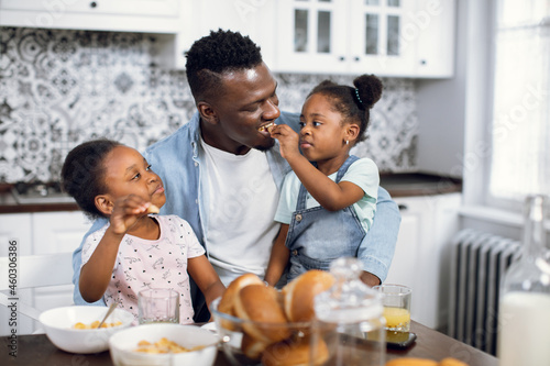 African american man and two cute daughters having breakfast together on bright kitchen. Happy family enjoying morning time at home. Concept of parenting and domestic life.