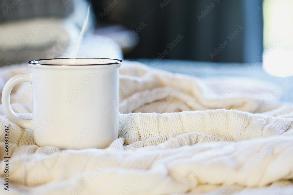 Hot steaming cup of coffee sitting on top a soft white knit blanket on a bed with stack of covers in background. Selective focus with extreme blurred foreground and background. Hot steaming cup of coffee sitting on top a soft white knit blanket on a bed with stack of covers in background. Selective focus with extreme blurred foreground and background.