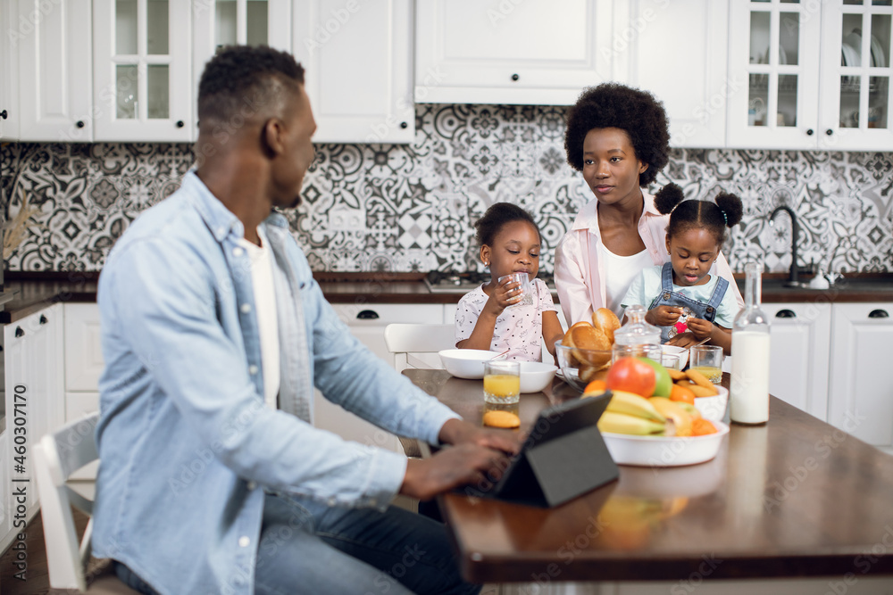 African american woman having breakfast with two cute daughters while husband working on digital tablet. Morning time of happy family on bright kitchen. African american woman having breakfast with two cute daughters while husband working on digital tablet. Morning time of happy family on bright kitchen.