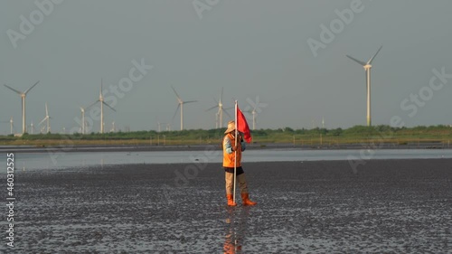 A coastal guard holding a red flag, standing still on the tidal flats with wind turbines spinning and rotating along the horizon at twilight, Gaomei wetland, Taichung, Taiwan. photo