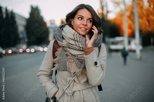 Smiling young brunette girl in a beige coat and a beige scarf walks through the streets of the autumn city and talking on a cell phone