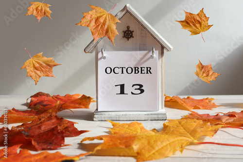 Calendar for October 13 : decorative house with numbers 13 and the name of the month in English, falling maple leaves , gray background, side view