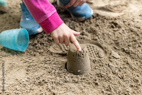 A picture of a child hands, wearing pink jacket and blue joggers, playing on sand while pointing on a sand model, made with a blue mold.