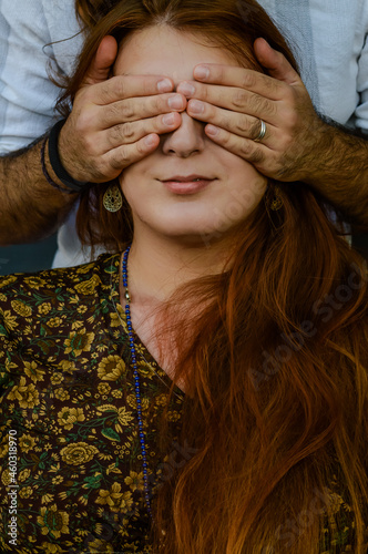 Portrait of a young red-haired woman with her eyes covered by the hands of a man