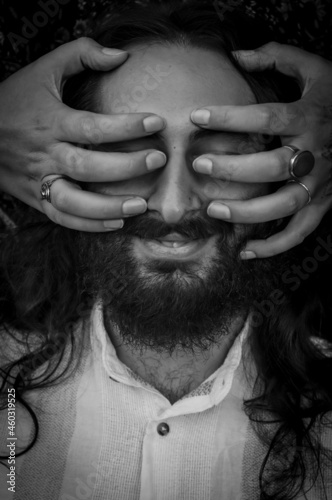 Fototapeta Naklejka Na Ścianę i Meble -  Black and white portrait of a young man with beard and long hair with eyes and face covered by a woman's hands