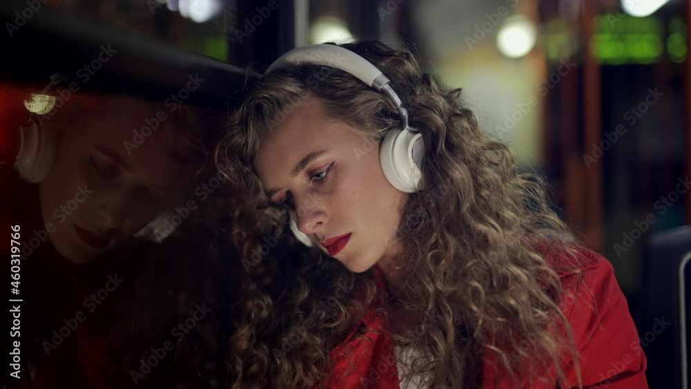 A depressed woman in headphones is riding in bus at night. Sad girl passenger is looking out window in modern tram. Depression or loss and problems or illness concept. City lights background.