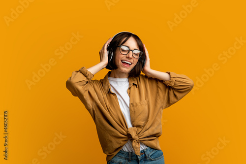 Carefree young lady in wireless headphones singing to her favorite music, enjoying playlist on yellow background © Prostock-studio