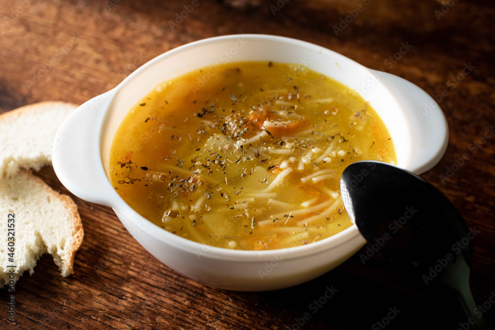 Chicken soup with noodles, potatoes, onions and carrots in a white deep plate with a black spoon and white bread on a wooden background. Chicken soup with noodles, potatoes, onions and carrots in a white deep plate with a black spoon and white bread on a wooden background.