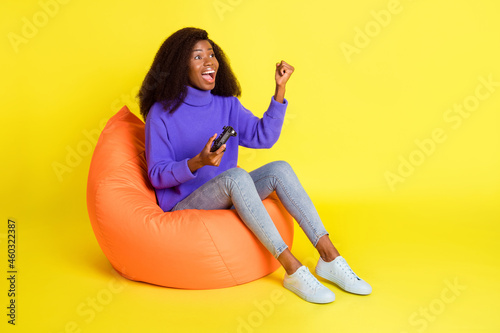 Photo of lady sit beanbag hold gamepad raise fist open mouth wear purple sweater jeans shoes isolated yellow color background