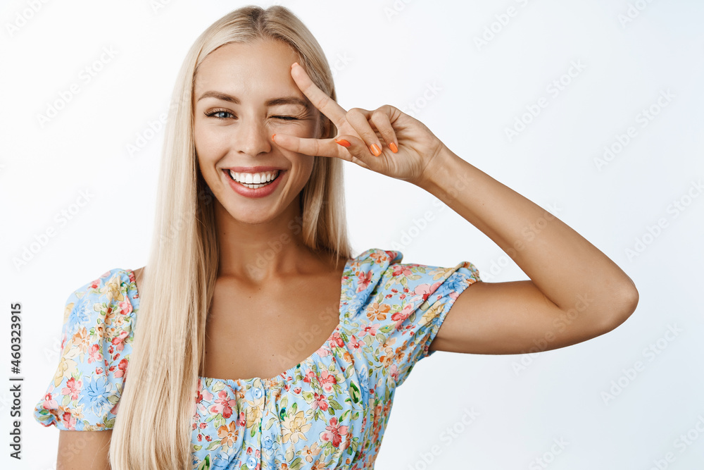Women beauty and skincare. Beautiful blond girl winking and smiling, showing peace v-sign salute and looking happy, standing in dress over white background Women beauty and skincare. Beautiful blond girl winking and smiling, showing peace v-sign salute and looking happy, standing in dress over white background