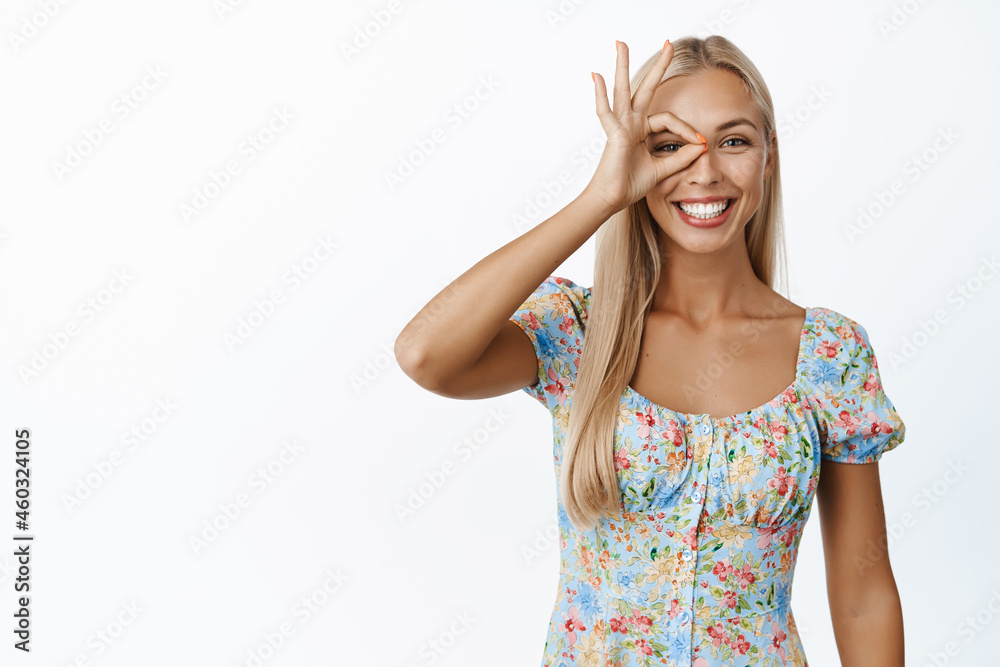Positive blond woman showing okay, zero gesture over eye, smiling, give positive answer, approve and like, standing over white background Positive blond woman showing okay, zero gesture over eye, smiling, give positive answer, approve and like, standing over white background