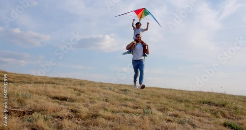 Father's day concept. Happy father and son running with a kite on blue sky. Cute happy boy sitting on dad shoulders launching kite together. 4k slow motion