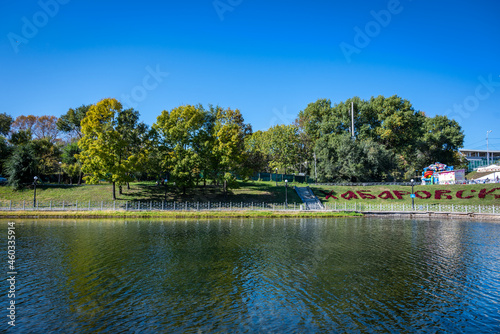 An autumn view of the public park in Khabarovsk (Russia) with blue clear sky, green and yellow trees, grass and a pond. On the grass there is an inscription "Khabarovsk" made of red flowers