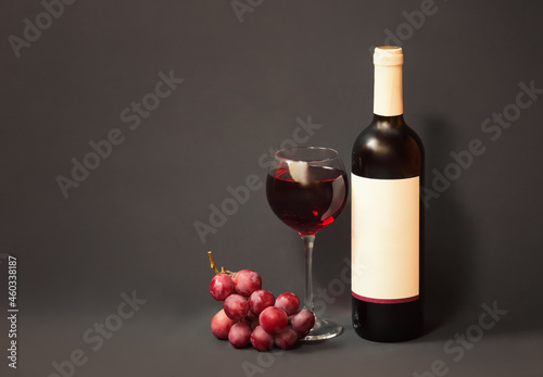 Bottle of red wine with white label, glass of red wine and purple grape on gray background.