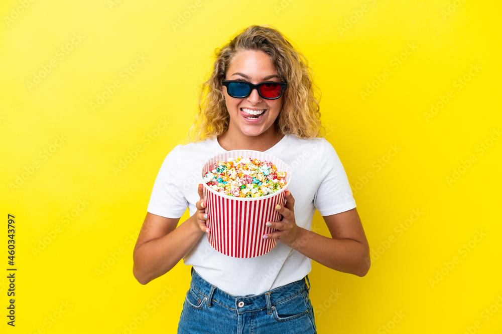 Girl with curly hair isolated on yellow background with 3d glasses and holding a big bucket of popcorns