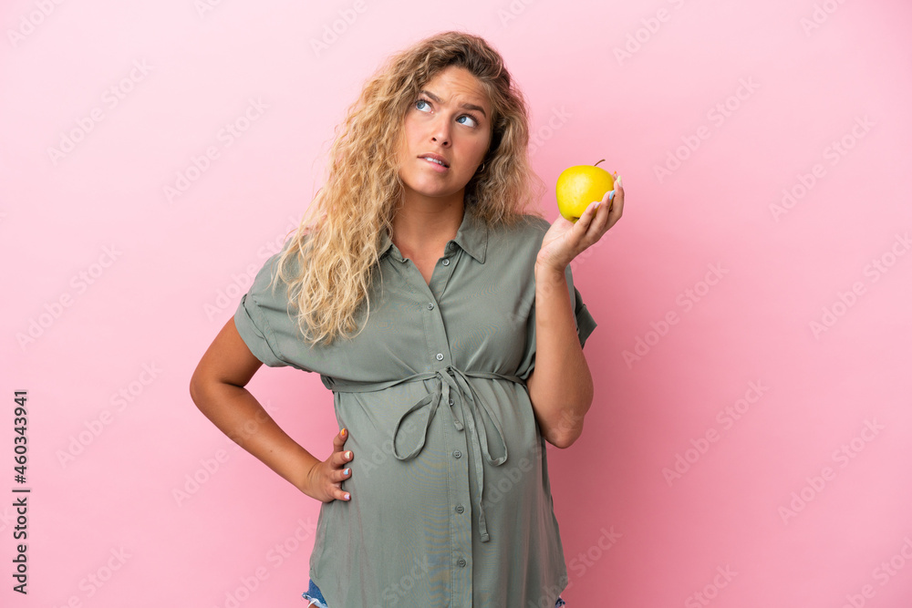 Girl with curly hair isolated on pink background pregnant and frustrated while holding an apple Girl with curly hair isolated on pink background pregnant and frustrated while holding an apple