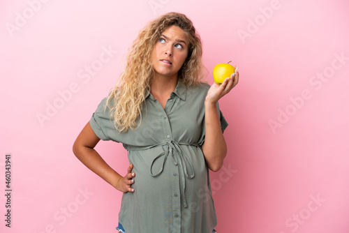 Girl with curly hair isolated on pink background pregnant and frustrated while holding an apple