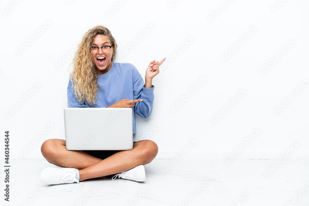 Girl with curly hair with a laptop sitting on the floor surprised and pointing side Girl with curly hair with a laptop sitting on the floor surprised and pointing side