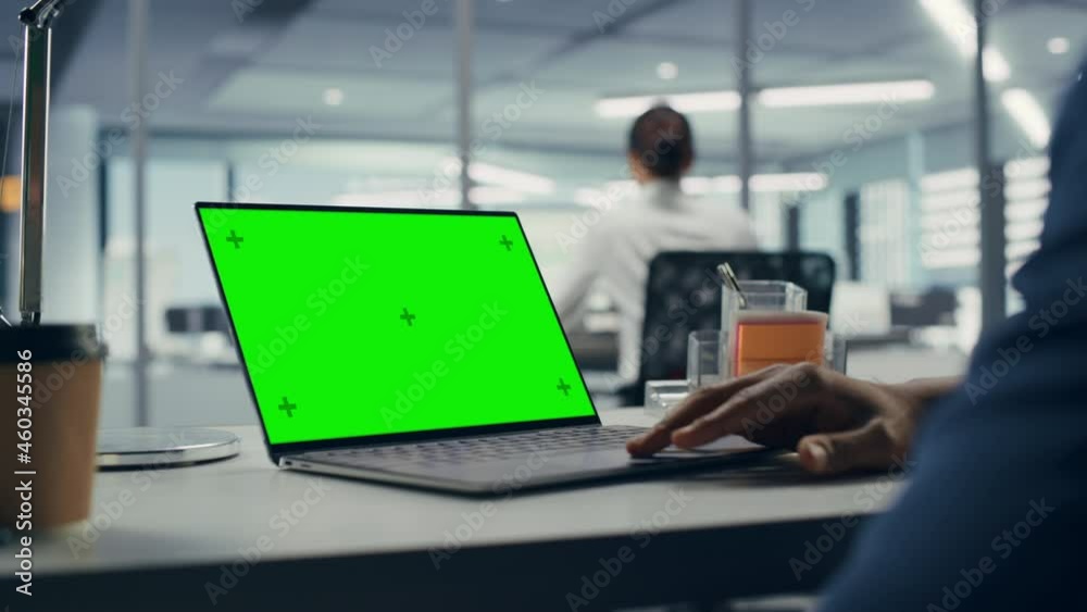 Successful Black Businessman Sitting at Desk Working on Green Screen Laptop Computer in Office. Anonymous Businessperson using Chroma Key Display. Stylish Bright Workplace. Over Shoulder Close-up