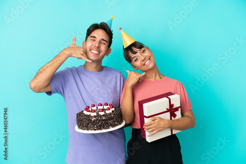Young mixed race couple holding birthday cake and present isolated on blue background making phone gesture. Call me back sign