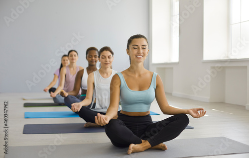 Diverse group of beautiful fit young women having meditation class at modern gym, sitting on yoga mats, doing lotus pose, smiling, relaxing, meditating with eyes closed and increasing energy levels
