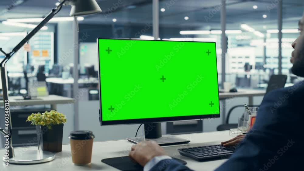Successful Black Businessman Sitting at Desk Working on Green Screen Laptop Computer in Office. African American Businessperson using Chroma Key Display. Over Shoulder Arc Shot Successful Black Businessman Sitting at Desk Working on Green Screen Laptop Computer in Office. African American Businessperson using Chroma Key Display. Over Shoulder Arc Shot