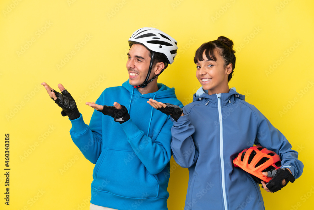 Young cyclist couple isolated on yellow background extending hands to the side for inviting to come Young cyclist couple isolated on yellow background extending hands to the side for inviting to come