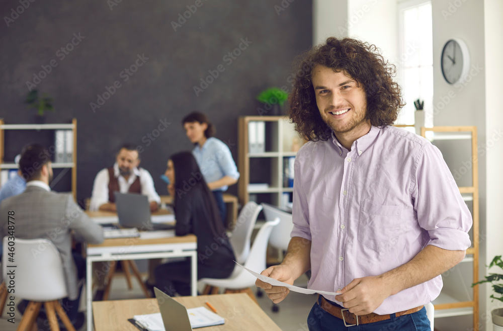 Happy handsome curly young man in casual shirt standing in modern office workplace, holding document, smiling and looking at camera. Portrait of successful confident business professional at work Happy handsome curly young man in casual shirt standing in modern office workplace, holding document, smiling and looking at camera. Portrait of successful confident business professional at work