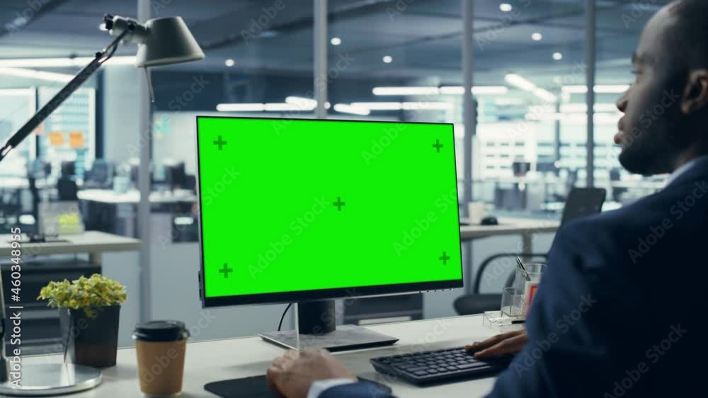 Successful Black Businessman Sitting at Desk Working on Green Screen Laptop Computer in Office. African American Businessperson using Chroma Key Display. Over Shoulder Zoom In