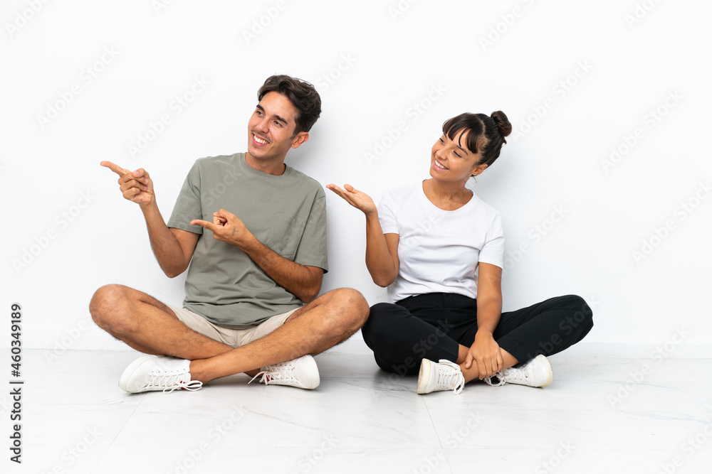 Young mixed race couple sitting on the floor isolated on white background pointing back and presenting a product Young mixed race couple sitting on the floor isolated on white background pointing back and presenting a product