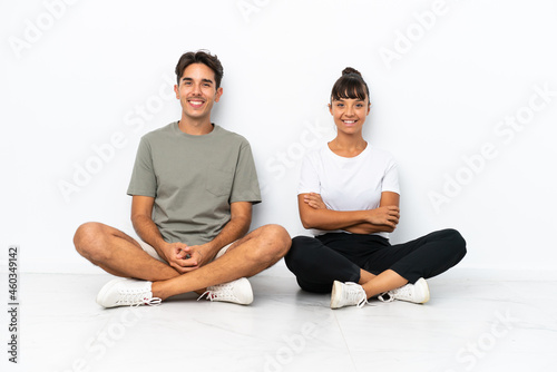 Young mixed race couple sitting on the floor isolated on white background keeping the arms crossed in frontal position