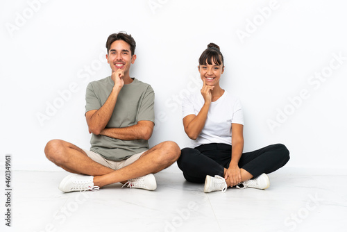 Young mixed race couple sitting on the floor isolated on white background smiling with a sweet expression